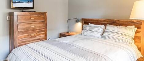 A bedroom with a wooden headboard, a bed with striped bedding, a nightstand with a lamp, and a TV mounted on a wooden cabinet.