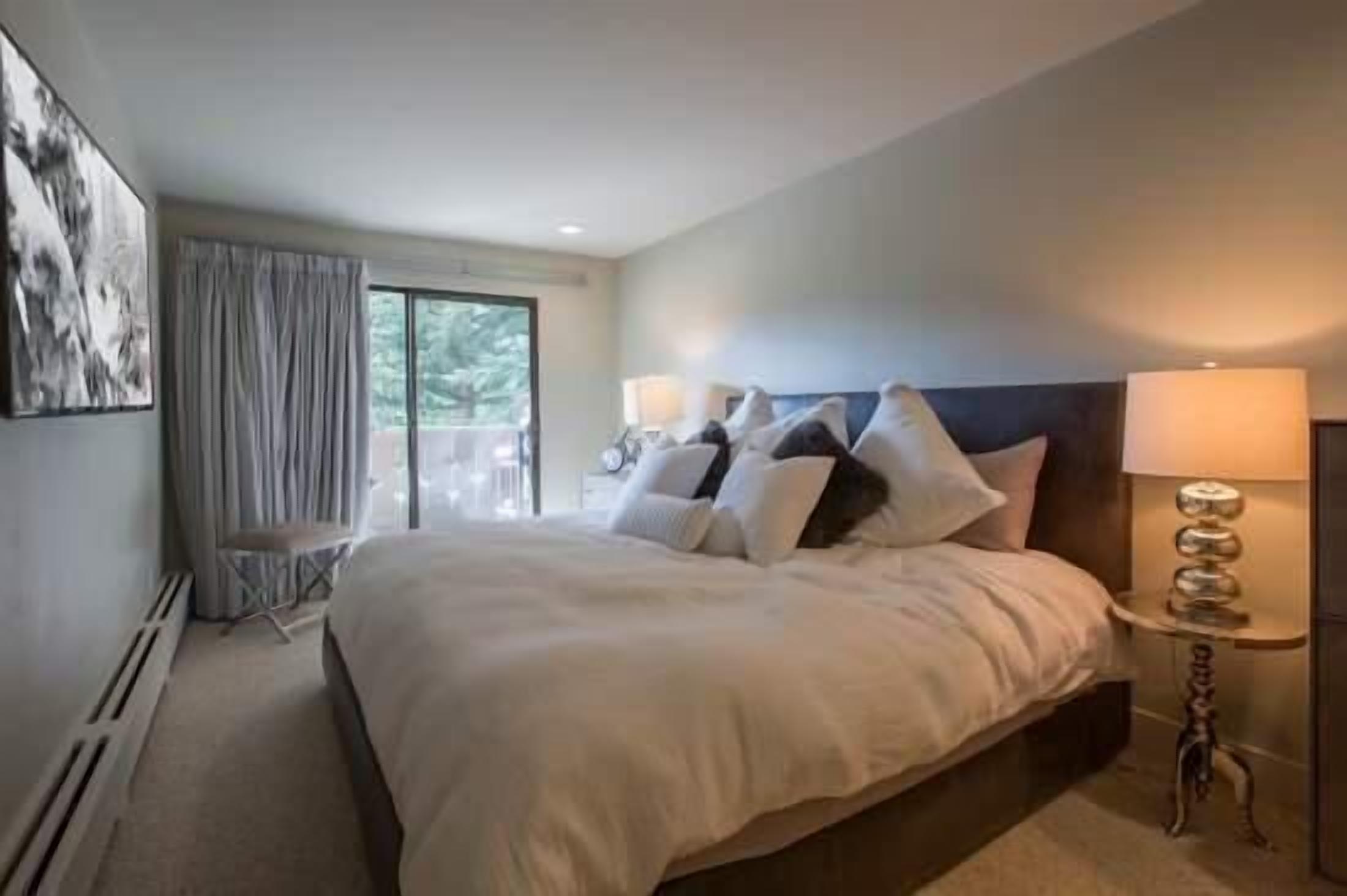 A modern hotel room with a large bed, two bedside lamps, a view of greenery through the window, and a small table with a decorative item.