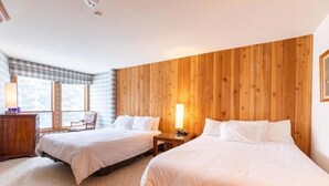 Een hotelkamer met twee bedden, een houten hoofdeinde, een raam met een geruit gordijn, een bureau met een lamp en een stoel.