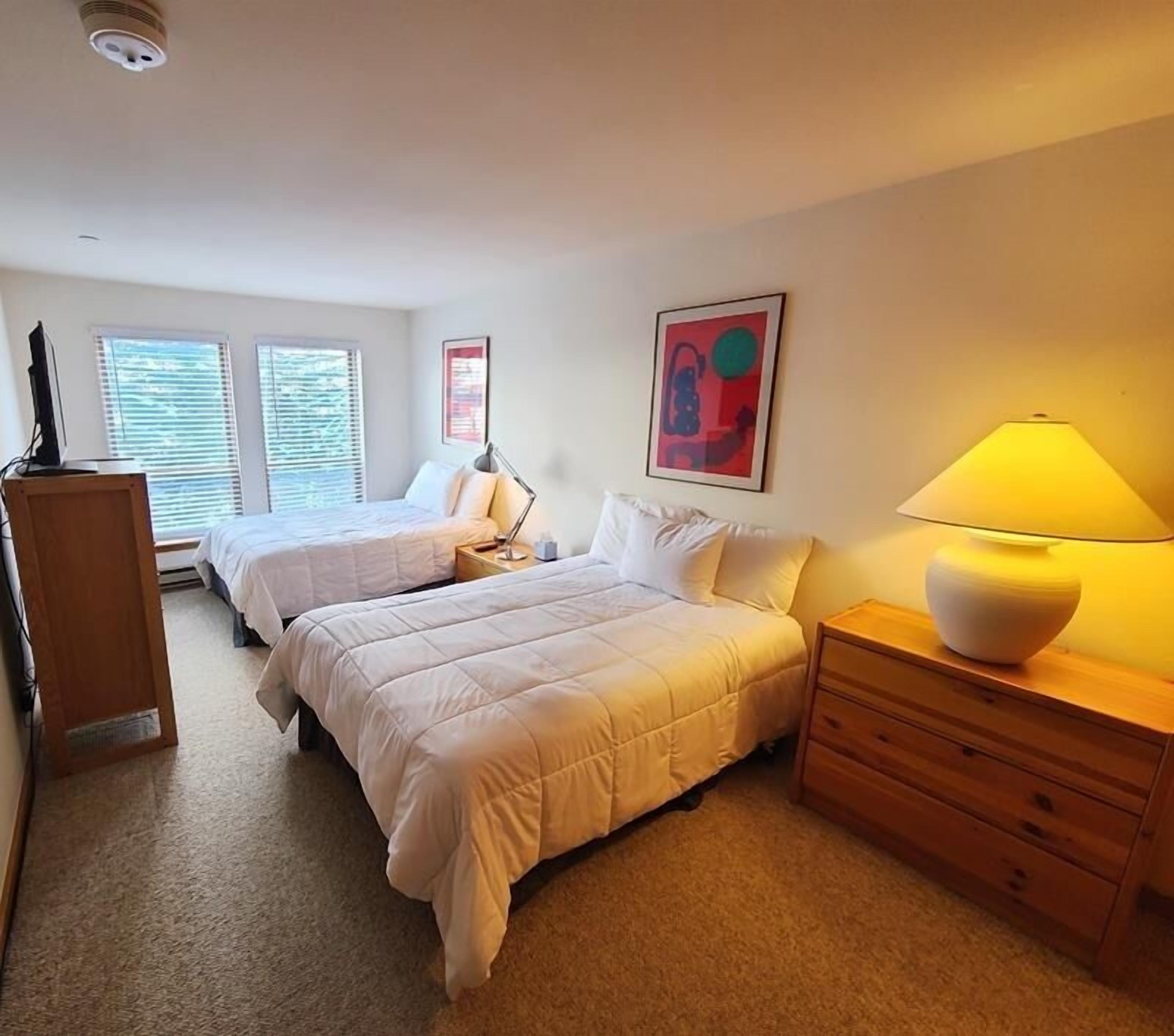 A bedroom with two beds, a desk, a lamp, and a painting on the wall.