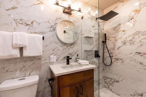 Un baño con pared de mármol, un espejo redondo, un lavamanos blanco, un armario de madera y una ducha.