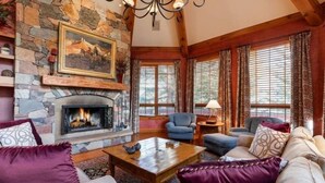 A cozy living room with a stone fireplace, comfortable seating, and large windows with curtains.