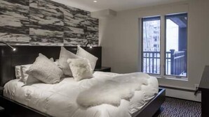 A modern bedroom with a large bed, a headboard with a textured design, and a window with a view of a fence. - Village Center Condos (Vail)