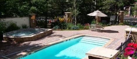 A rectangular swimming pool with a hot tub on one side, surrounded by a brick patio and lush greenery.