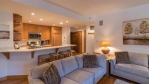 Una cocina moderna con armarios de madera, una isla central y taburetes de bar. Hay una zona de comedor con un sofá y una lámpara.