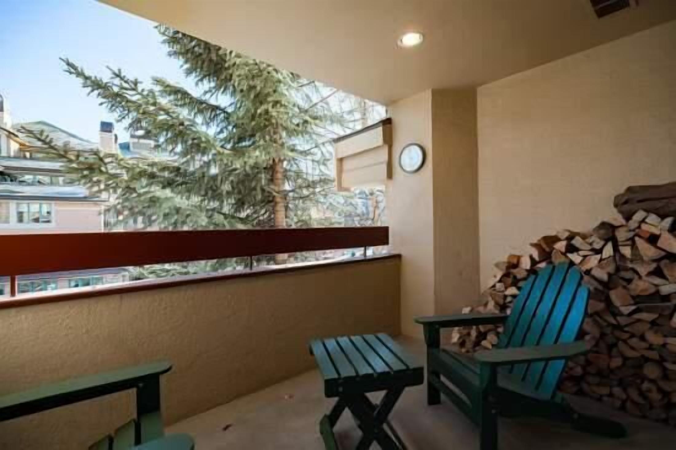 A balcony with a wooden table and chair, a clock on the wall, and a stack of firewood.