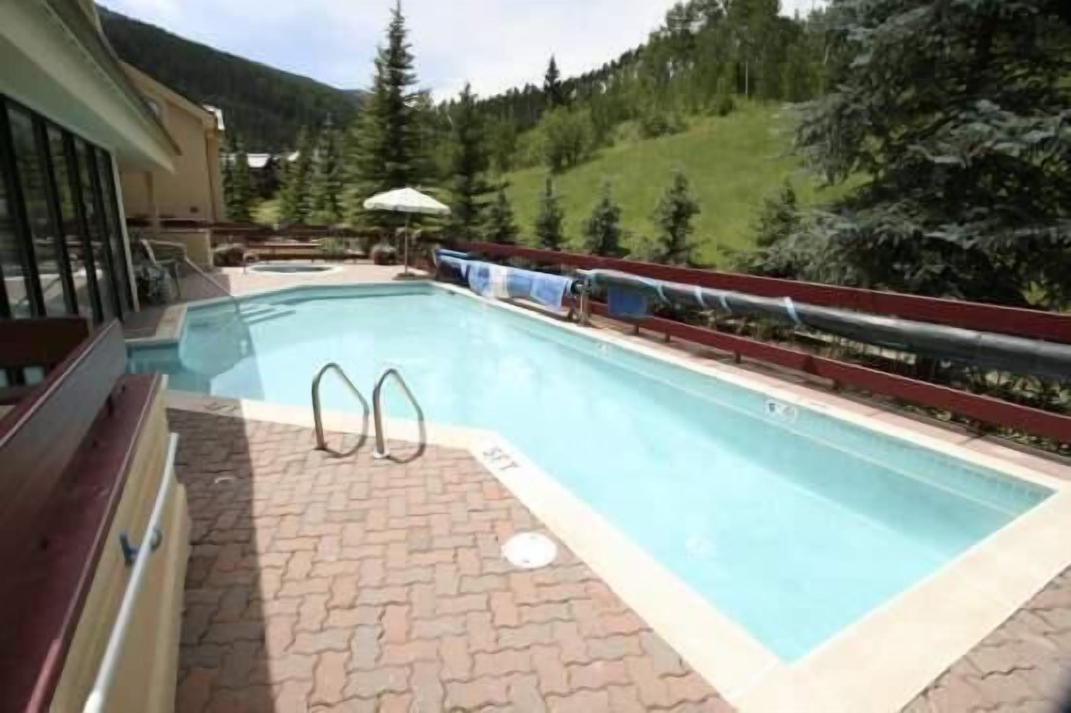 Una piscina con cubierta de madera, barandilla roja y montañas al fondo.
