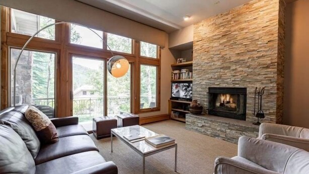 A living room with a stone fireplace, large windows, a sofa, and a coffee table. - Ski-in Ski-out Townhome in Beaver Creek by RedAwning (Avon)