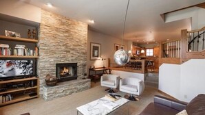 A modern living room with a stone fireplace, a television, and a dining area with a staircase. - Ski-in Ski-out Townhome in Beaver Creek by RedAwning (Avon)