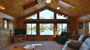 A cabin with a large window overlooking a lake, a leather sofa, a wooden coffee table, and a television on a cabinet.