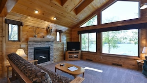 A cozy cabin interior with a fireplace, a television, a sofa, and large windows offering a view of the outdoors.