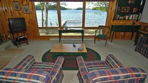 A cozy cabin interior with a large window overlooking a lake, plaid furniture, and a bookshelf. - Ranch-Style Seidls Home Feet away to Lakeshore by RedAwning (Cable)