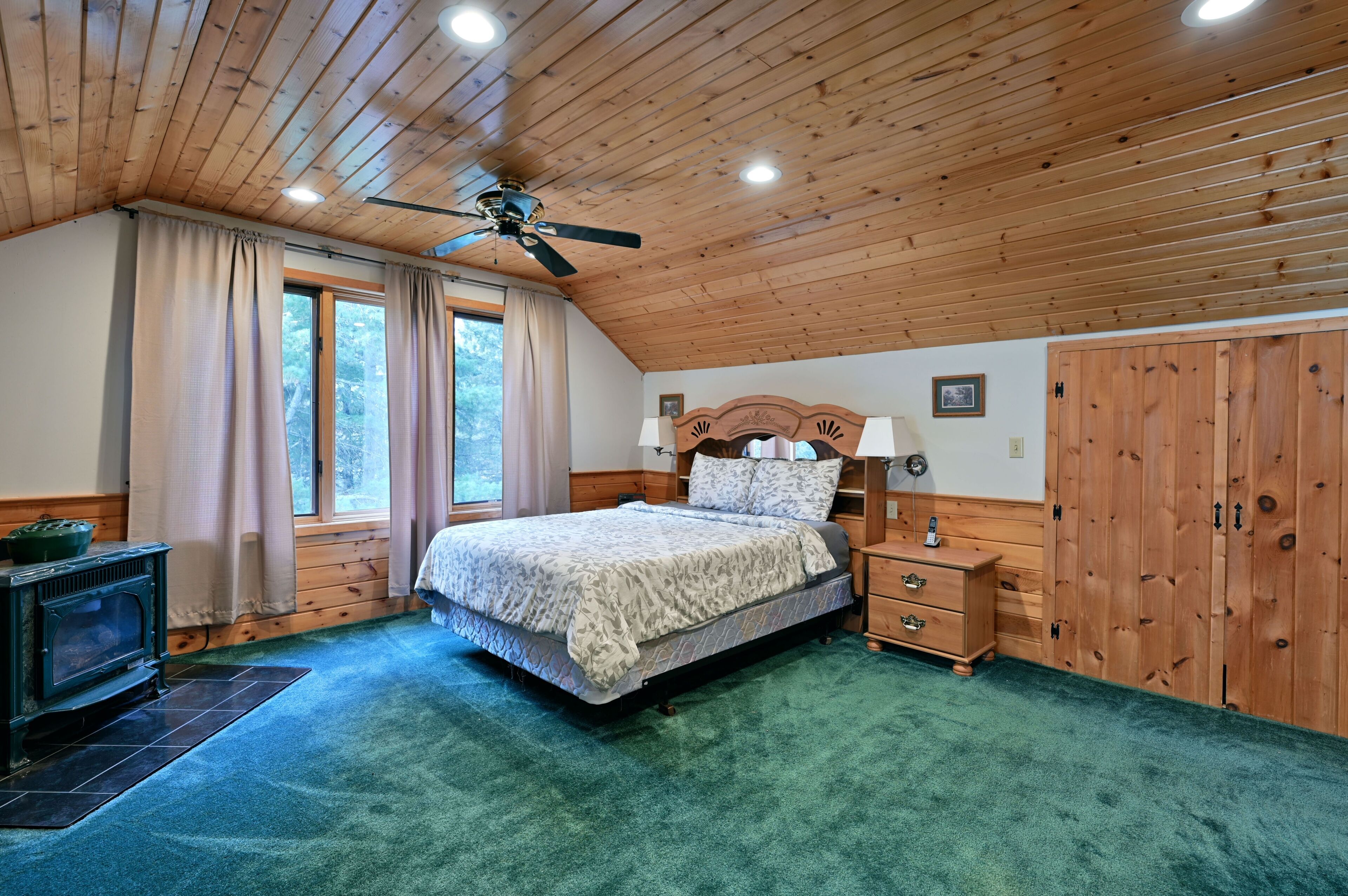 A bedroom with a wooden ceiling, a bed with a headboard, a nightstand, a dresser, and a window with curtains.