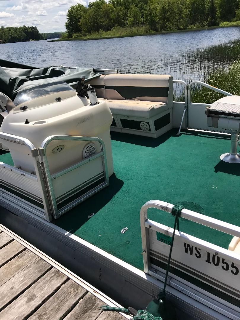 Un bote amarrado en un muelle, con cubierta verde, cabina blanca y una cama doble, además de una pequeña mesa.