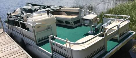A pontoon boat docked at a wooden pier with a green deck, a motor, and seating areas.