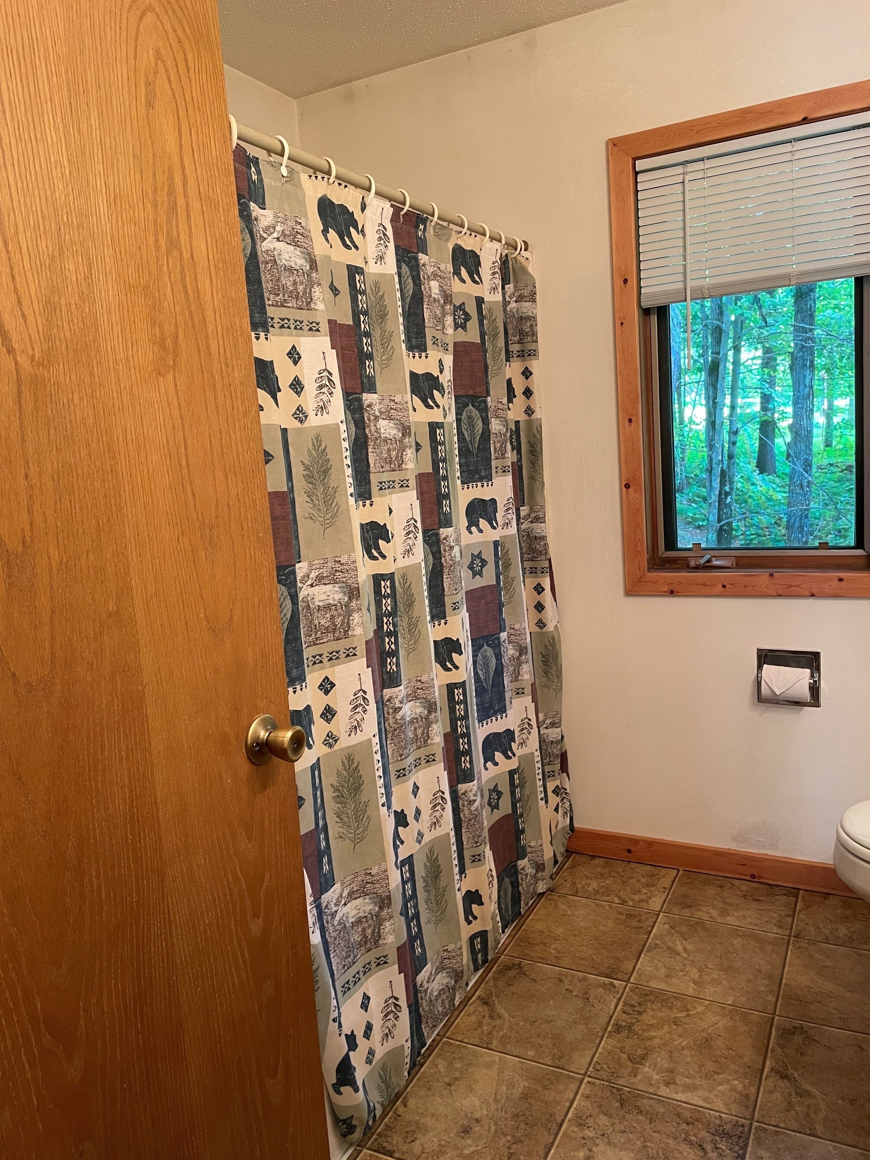 A bathroom with a shower curtain featuring a bear and leaf pattern, a window with blinds, and a toilet with a paper towel holder.