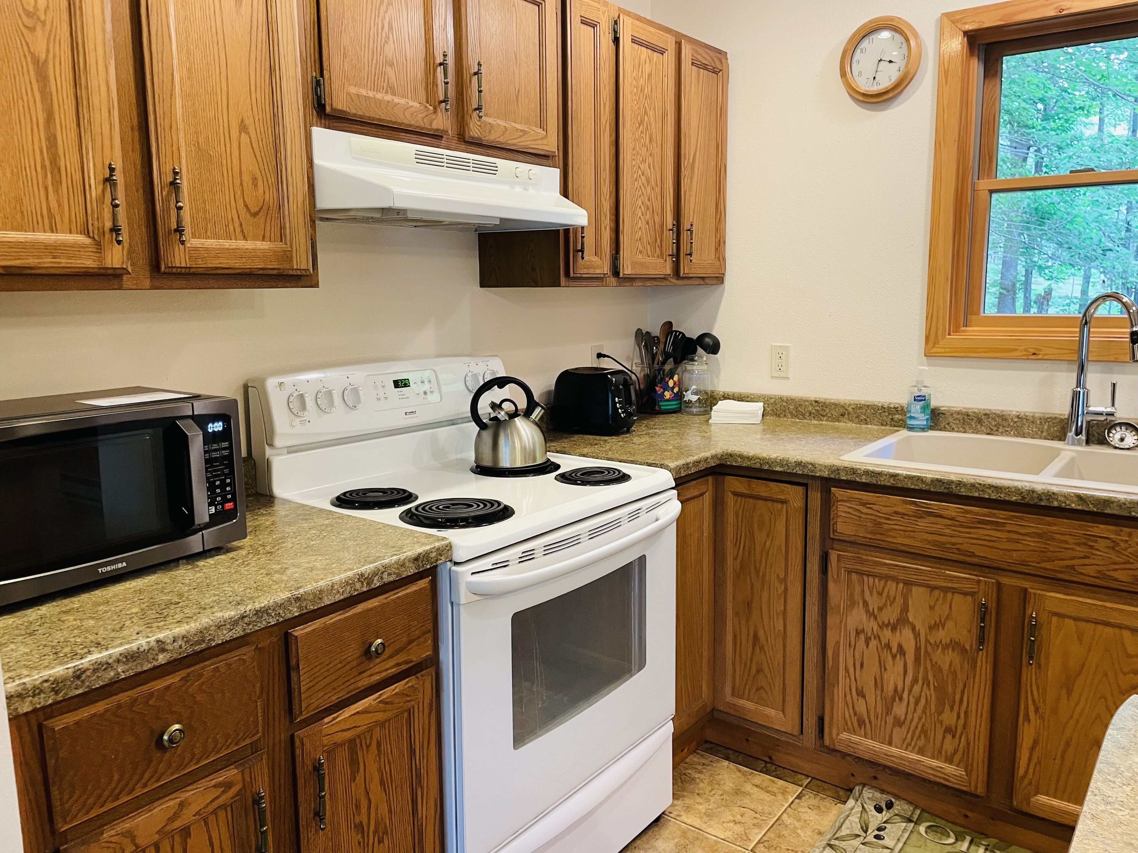 A kitchen with wooden cabinets, a microwave, stove, and a window with a view of trees.