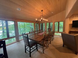 Un comedor amplio con una larga mesa de madera y sillas, ventanas grandes que ofrecen vistas al bosque, y una araña colgando del techo.