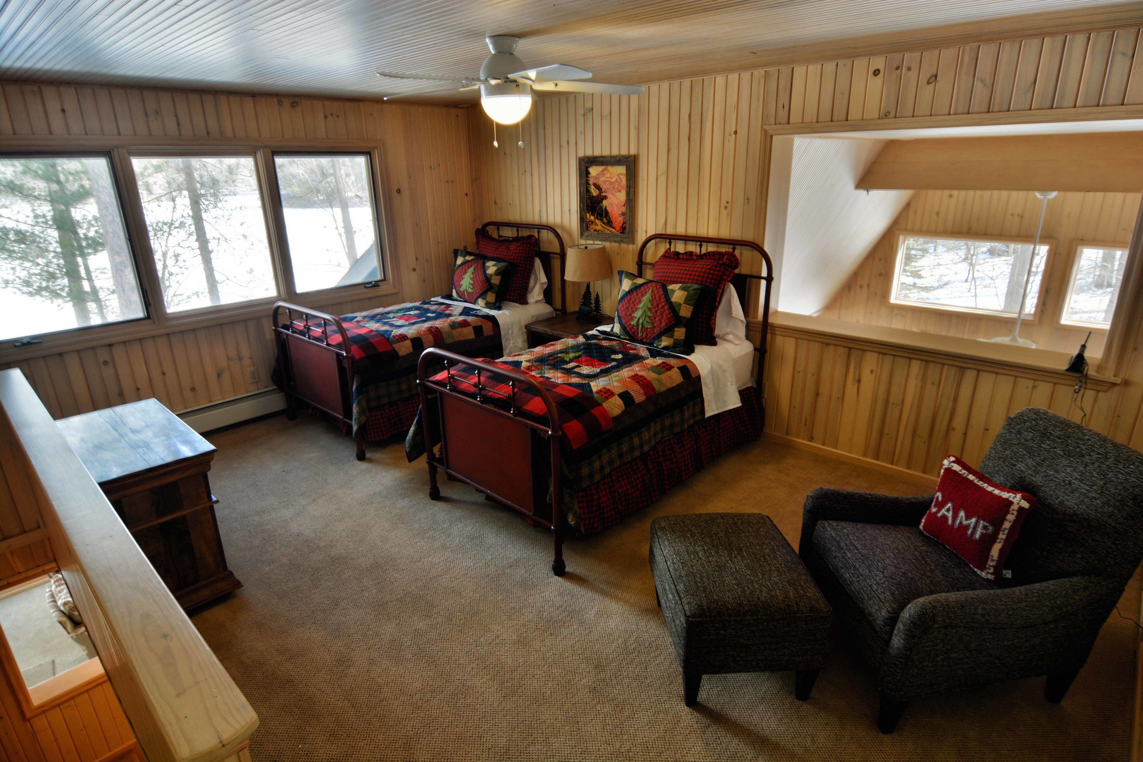 A cozy cabin room with two beds, a chair, a bench, and a wooden desk.