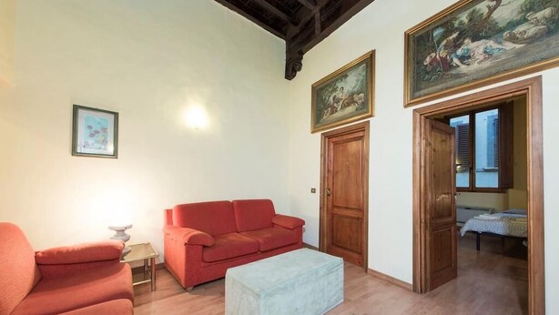 A living room with a red sofa, a light-colored ottoman, a small table with a lamp, and a door leading to another room. - Sdrucciolo Pitti (Florence)