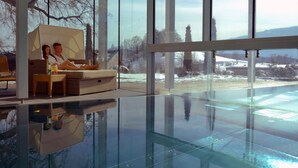A modern indoor pool area with glass walls, a lounge area with wicker furniture, and a view of a snowy landscape. - Das Moerisch (Seeboden)