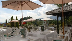An outdoor dining area with white tables and chairs, beige umbrellas, and a scenic mountain backdrop. - Das Moerisch (Seeboden)