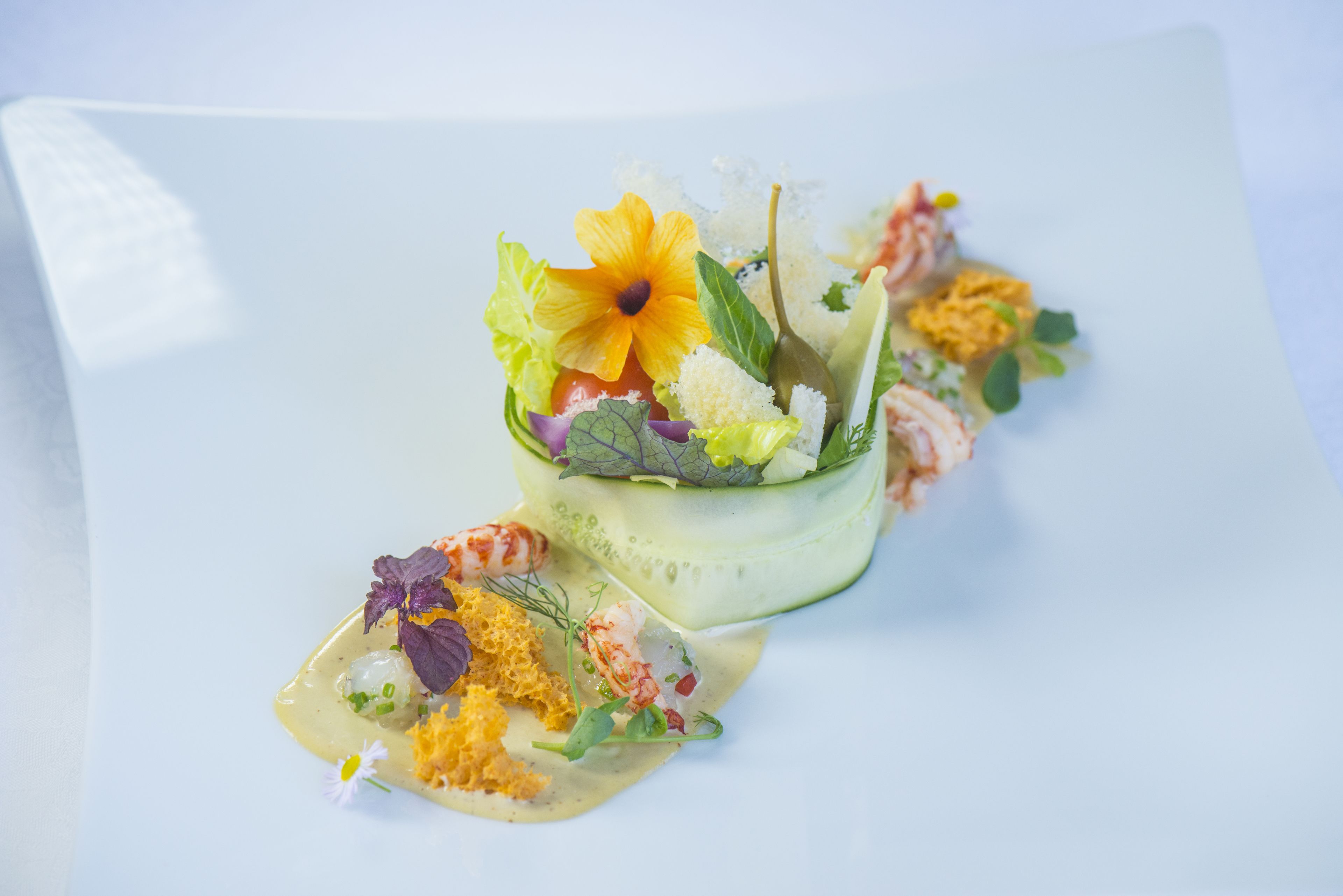 Een witte schaal met een zeevruchtengerecht, een plak komkommer en een decoratieve bloem.