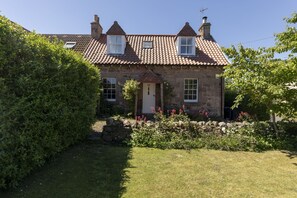 Een stenen huis met pannendak, een voordeur en een kleine tuin met bloemen en struiken.
