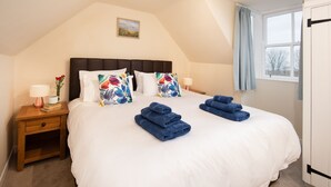 A neatly made bed with white bedding and blue towels, a nightstand with a lamp and books, and a window with curtains. - WINDYRIG, Cockburnspath - sleeps 4 in 2 bedrooms (Cockburnspath)
