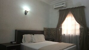 A hotel room with a bed, a nightstand, a wall-mounted air conditioner, and a window with curtains. - Rila Muam Castle Hotel (Kaduna)