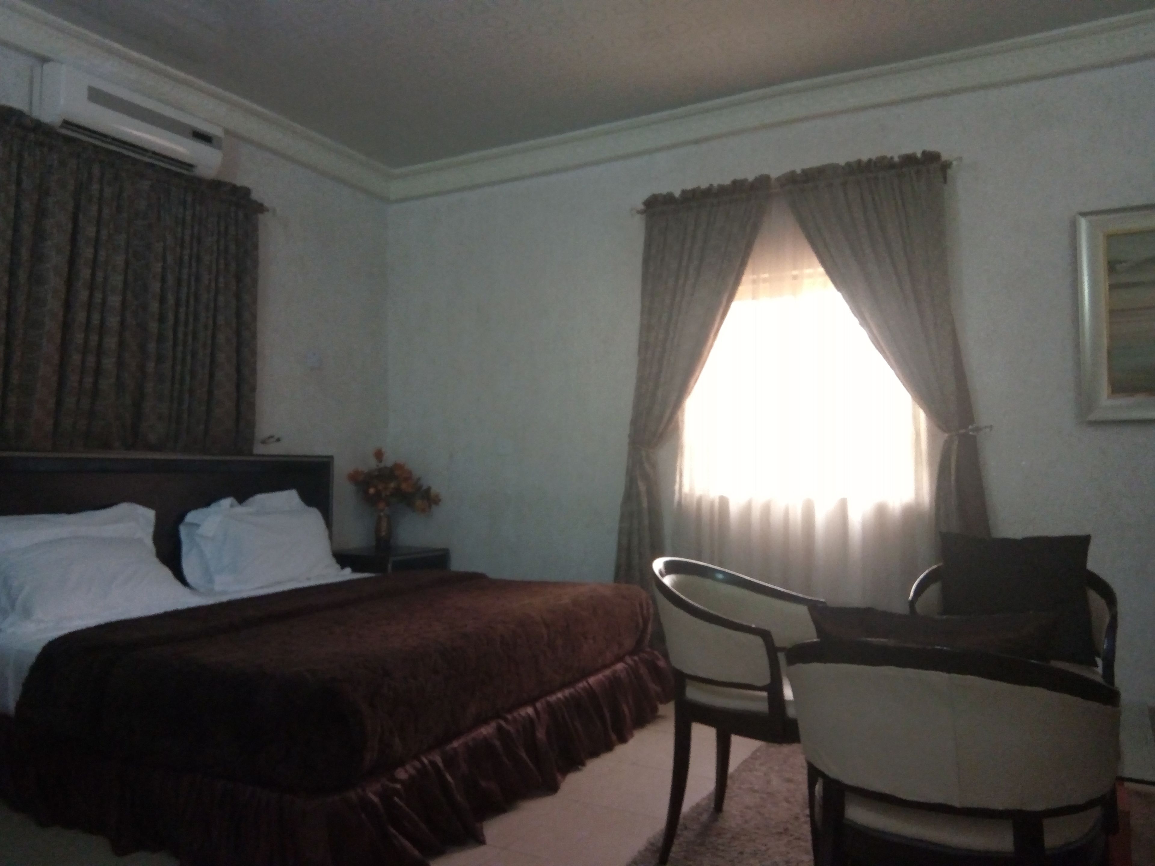 A hotel room with a bed, a desk, a chair, a window with curtains, and an air conditioning unit.