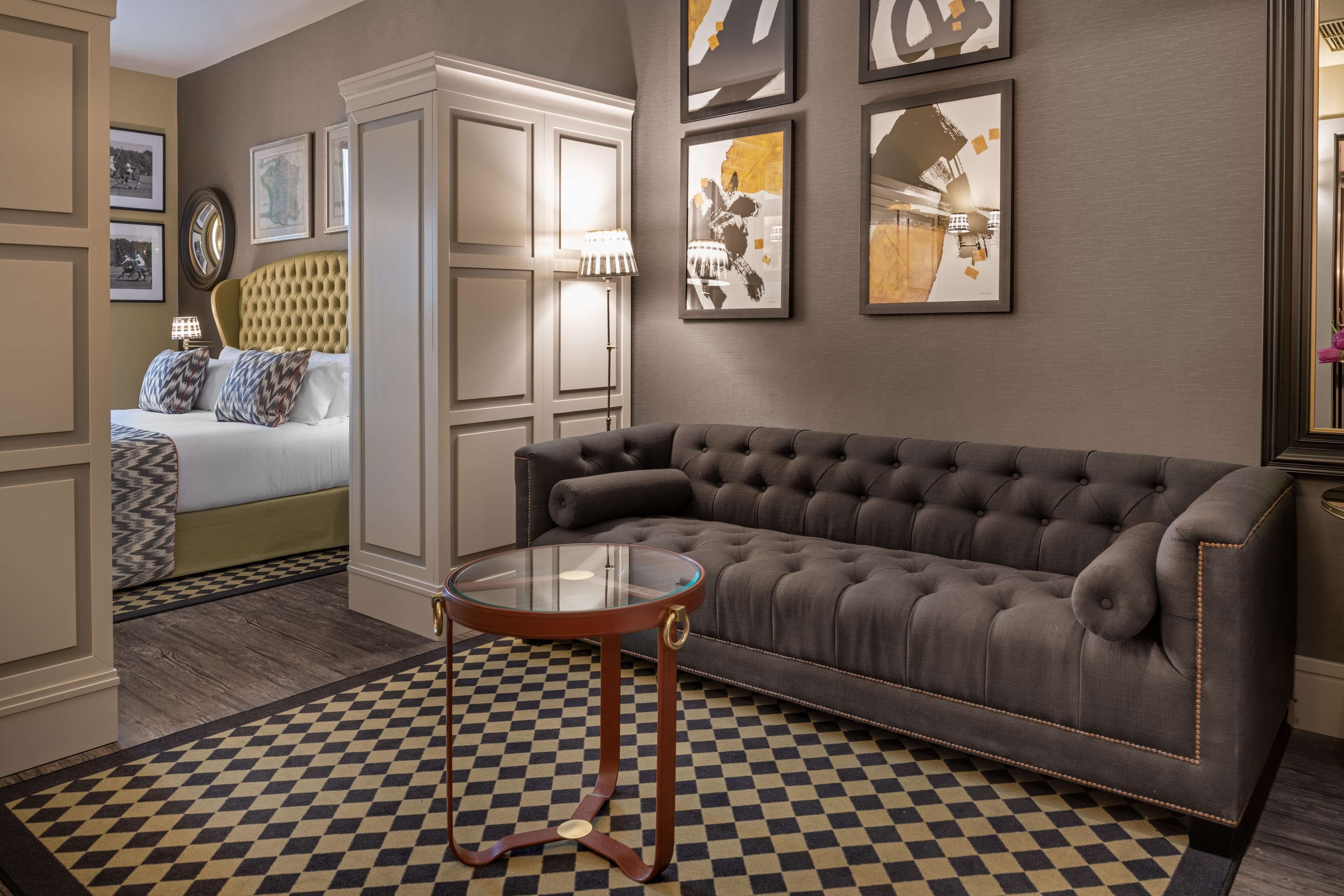 A hotel room with a checkered floor, a tufted sofa, a glass table, a bed with patterned pillows, and framed artwork on the walls.