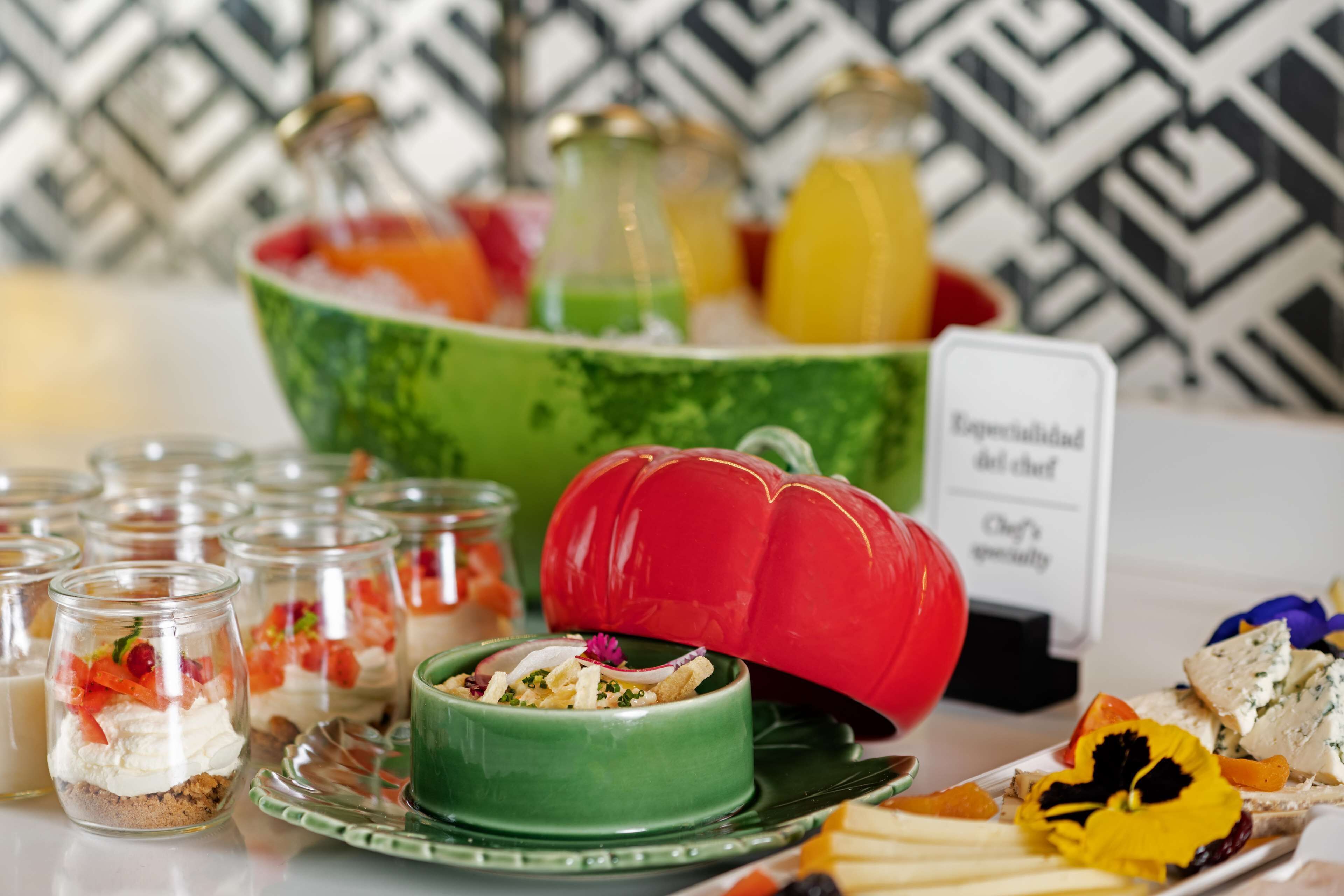 Una mesa de buffet con diversos alimentos, incluyendo un tazón grande de fruta, un pimiento rojo y varios recipientes de vidrio pequeños con postres.
