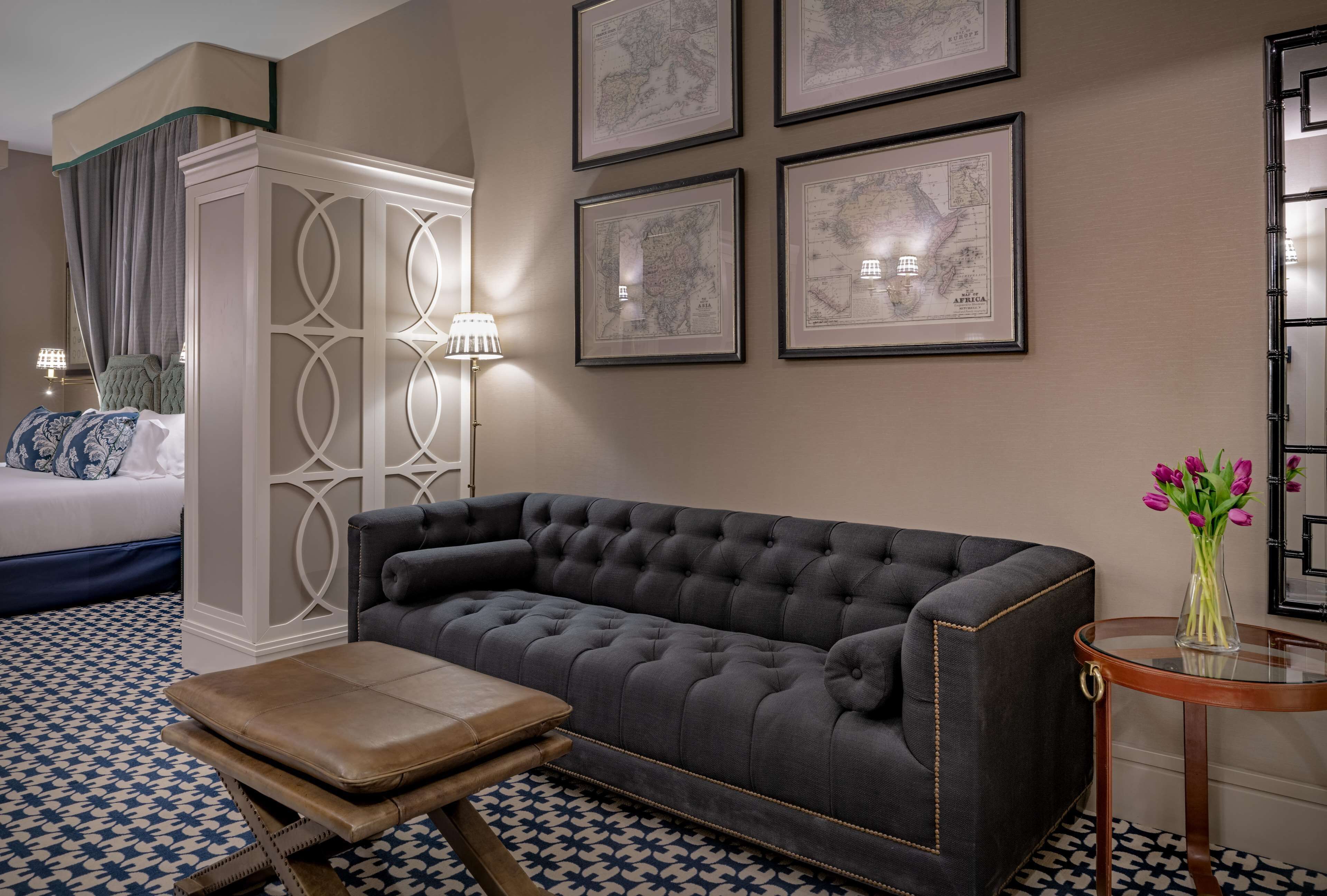 Una habitación de hotel moderna con un sofá gris con capitoné, un banco de madera, una pequeña mesa redonda con un jarrón de flores y una cama con colcha de diseño azul y blanco.