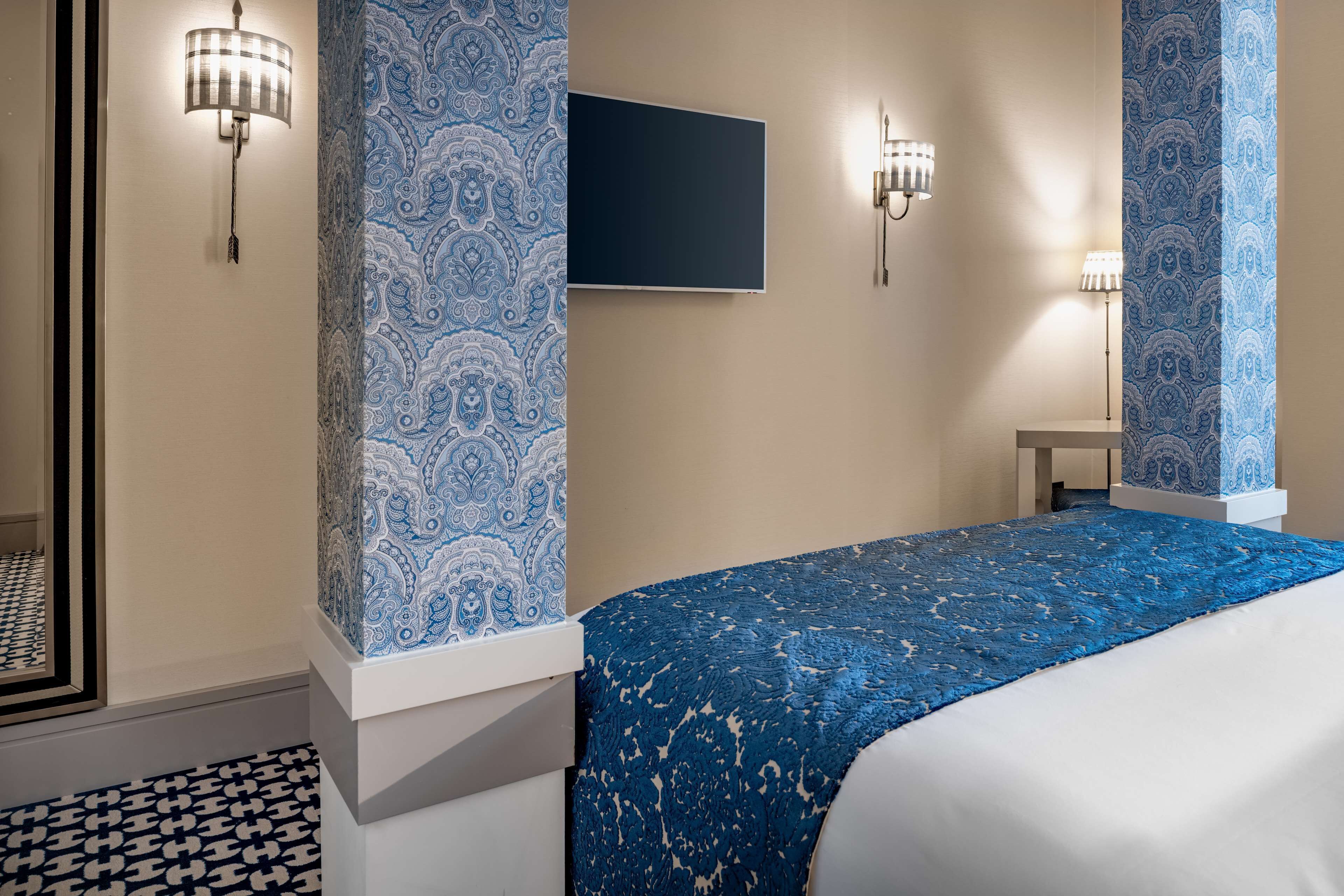 Habitación de hotel con cama, papel tapiz estampado, luces fijadas a la pared y televisor de pantalla plana.