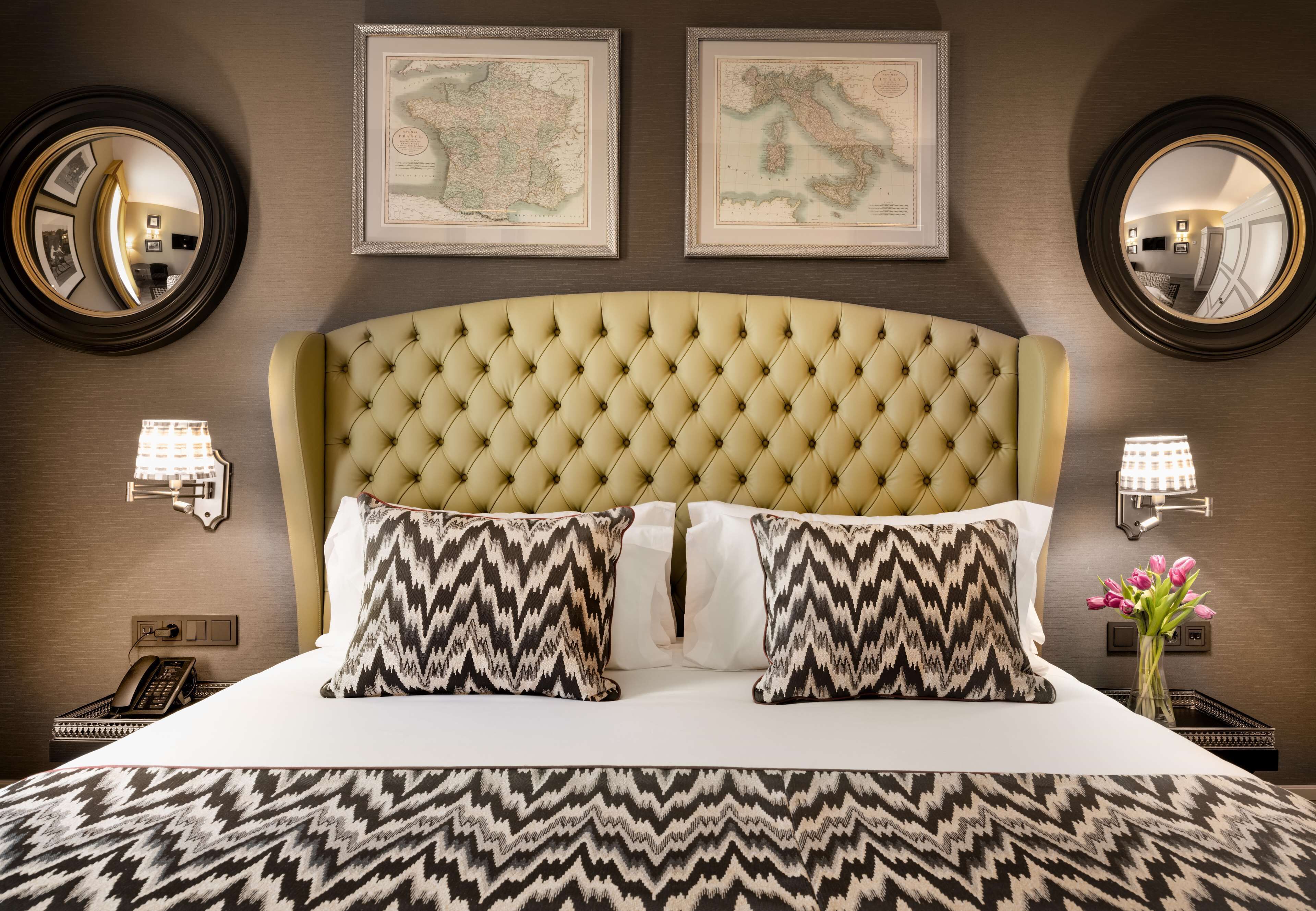 Une chambre d’hôtel avec une tête de lit capitonnée, deux cartes encadrées accrochées au mur, un lit agrémenté d’oreillers à motifs, des lampes de chevet et un vase rempli de fleurs.