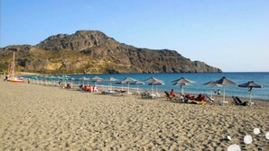 Una playa con sombrillas y sillas reclinables, una montaña rocosa al fondo y un cielo despejado.