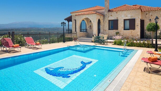 Une piscine décorée avec un motif de dauphin, des chaises longues et un bâtiment en pierre surmonté d’un toit en tuiles.