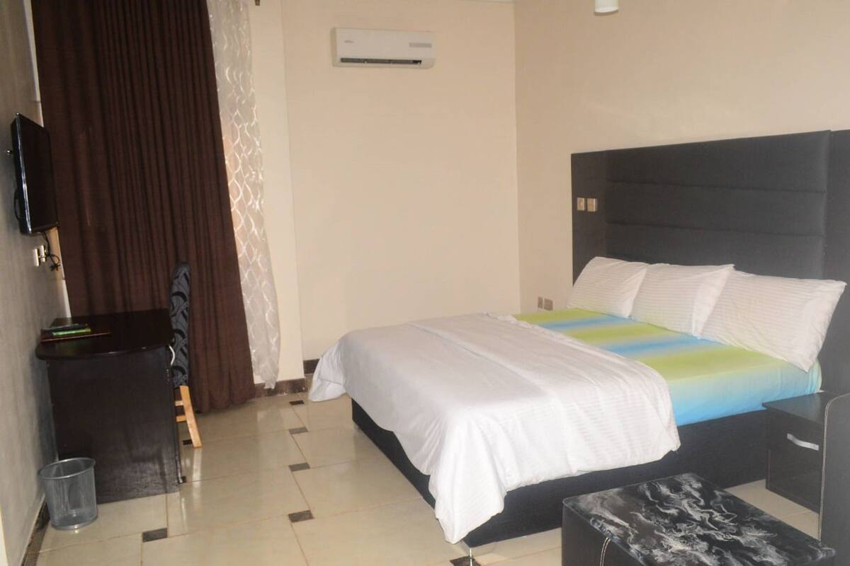 A hotel room with a bed, a desk, a chair, an air conditioning unit, and a trash bin.