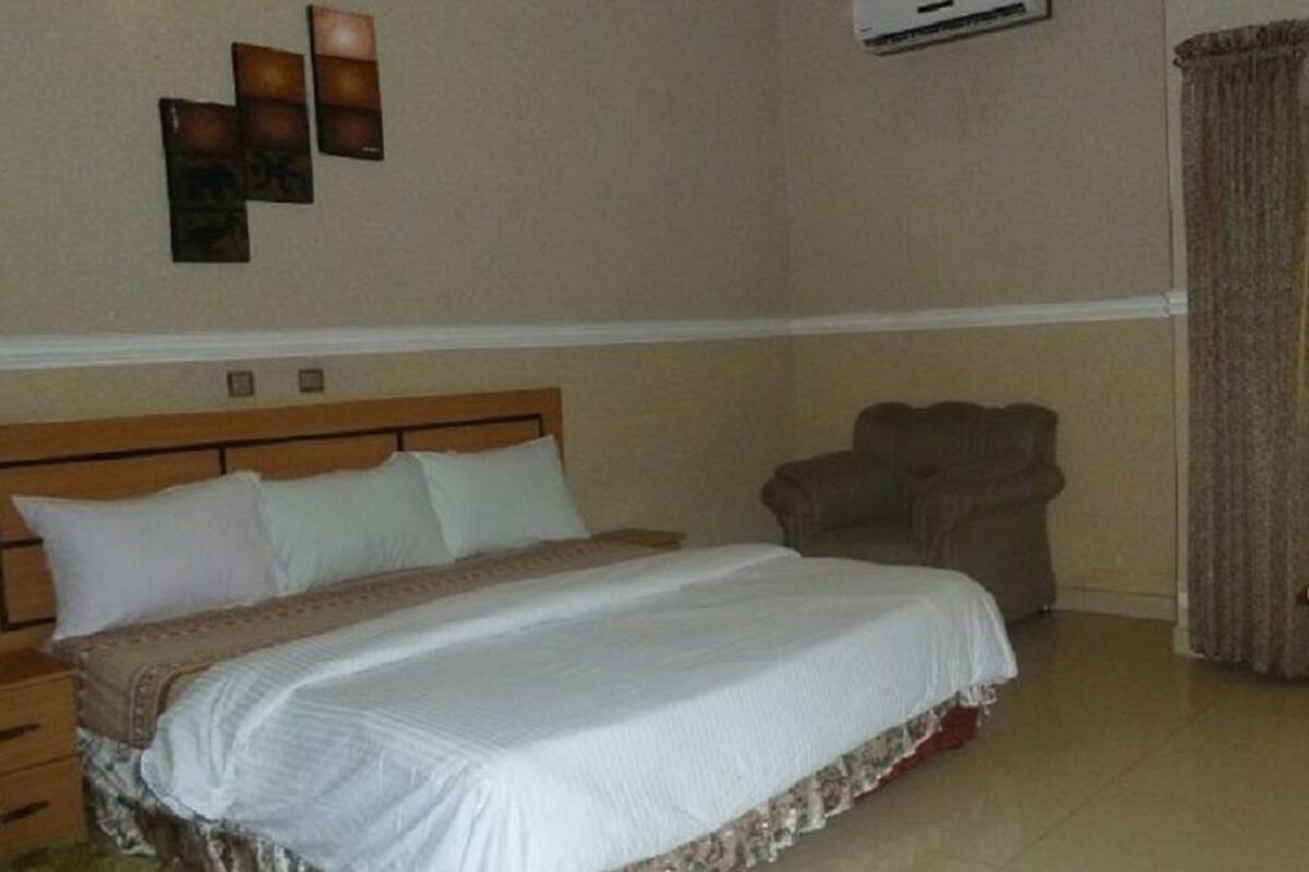A hotel room with a large bed, a wooden headboard, a chair, and a wall-mounted air conditioner.