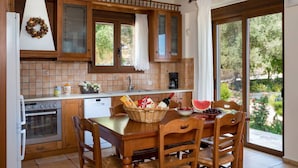 A kitchen with wooden cabinets, a dining table with chairs, a window with a view of greenery, and a chandelier. - Family Villa w/Pool, Billiard ,Ping Pong, BBQ area (Chania)