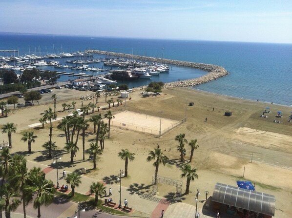 A marina with boats docked, a sandy beach with palm trees, and a clear view of the sea. - Finikoudes Luxury Apartments (Larnaca)