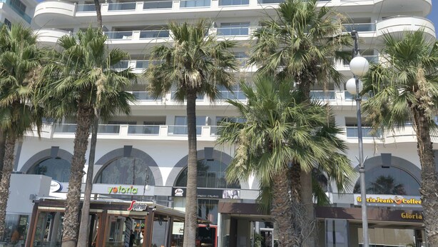 Un hotel a più piani con balconi, palme e negozi, tra cui un caffè.