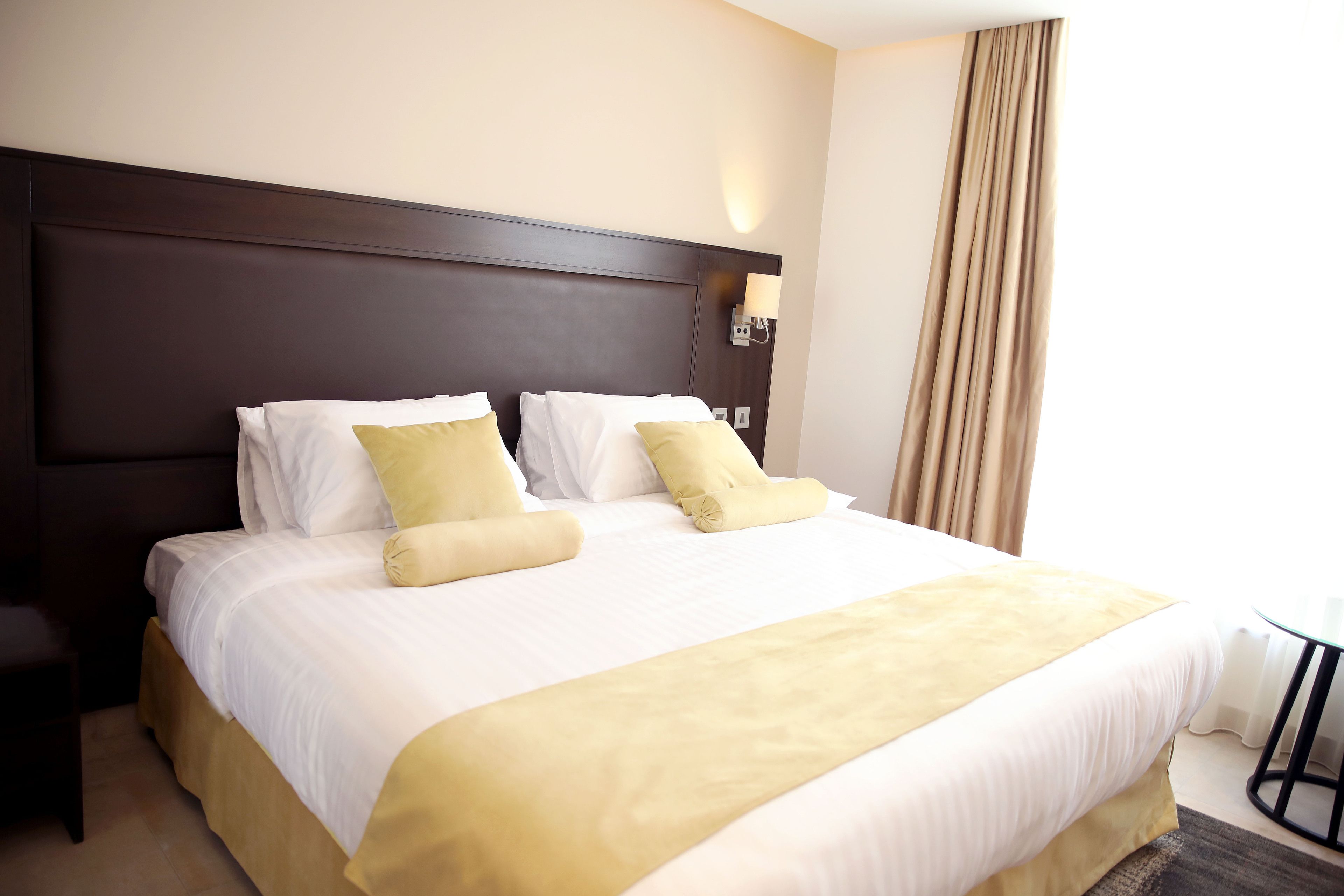 Una habitación de hotel con una cama grande, cabecero, mesita de noche y ventana con cortinas.