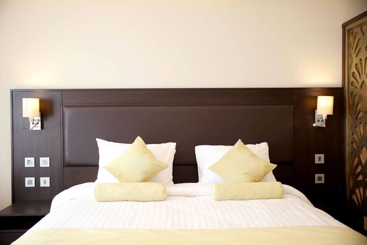 A hotel room with a dark wood headboard, two bedside lamps, a bed with white bedding and two beige pillows, and a decorative wall panel.