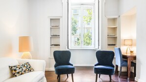 A living room with a white sofa, two blue armchairs, a wooden dining table, and a window with a view of greenery. - easyhomes - Majno (Milan)