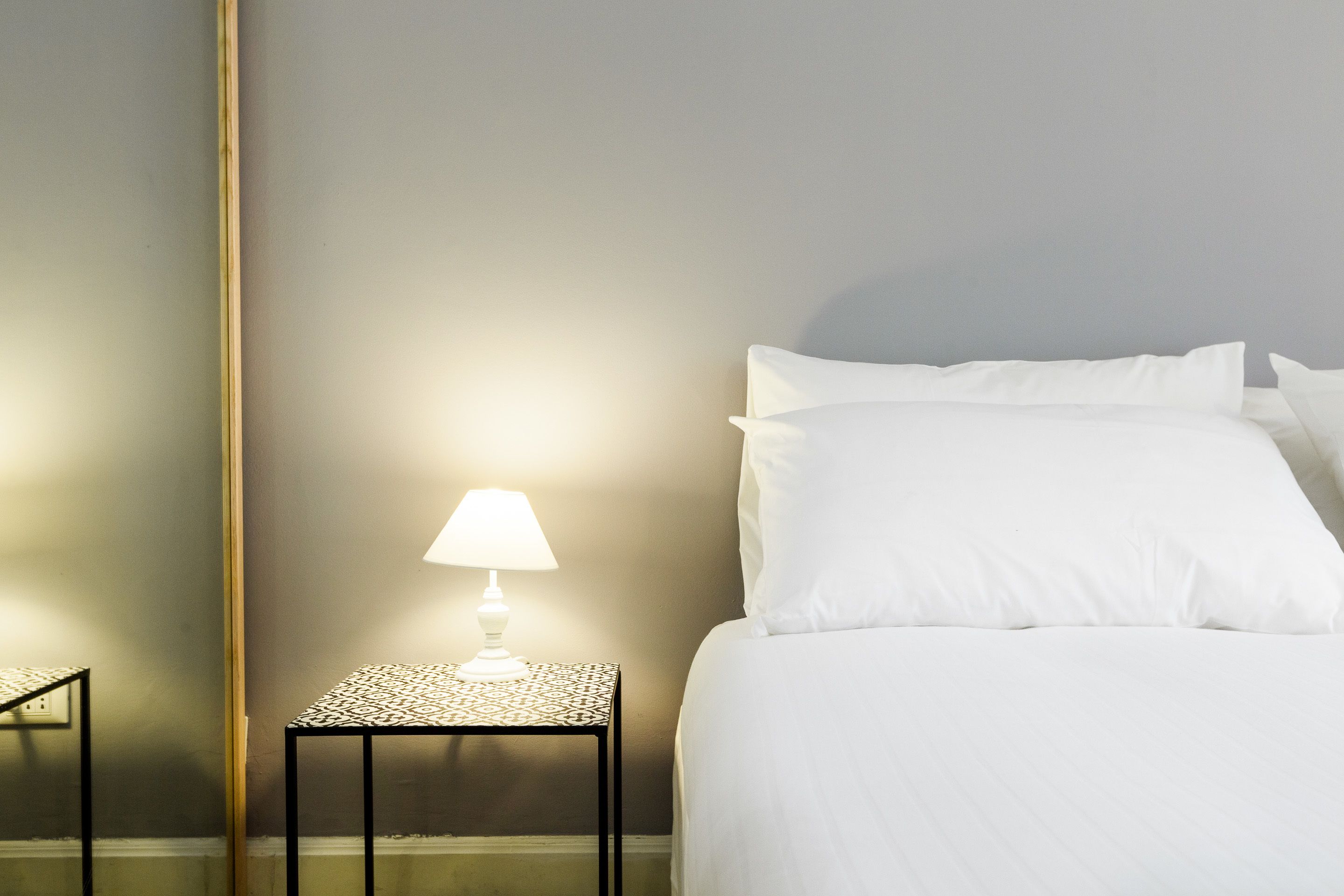 A neatly made bed with white linens, a small bedside table with a lamp, and a wall with a vertical wooden element.
