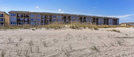 Een strandhotel met meerdere balkons, een zandstrand op de voorgrond en heldere lucht.