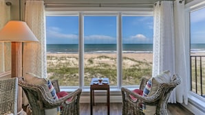 Ein Zimmer mit Korbstühlen, einem Tisch mit Tellern und Blick auf Strand und Meer durch große Fenster.