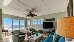 Un salón con vistas al océano, un ventilador de techo, un televisor y un letrero que dice 'She sells seashells by the sea shore'.